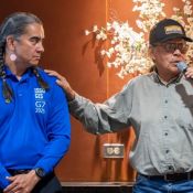 An Indigenous man speaks into a microphone while another wearing a G7 branded shirt listens.