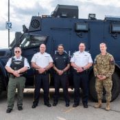 Five men in uniforms from different law enforcement and defence agencies stand in front of an RCMP vehicle.