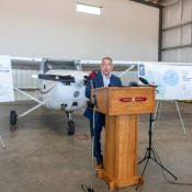Man in a suit speaks behind a podium in an airplane hanger. A small plane and posters are displayed in the background.