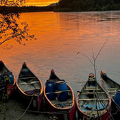 Five canoes are docked on the shore of a river. A deep orange sunset reflects in the calm water with a view of mountains are in the background.