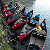 Seven red canoes packed with fuel and waterproof containers are docked on the shore of a river. 