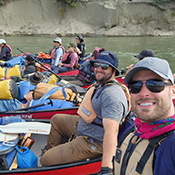 Canoe paddlers line up in their canoes along the river's shore. The canoers are unloading the canoes which are packed with supplies. 