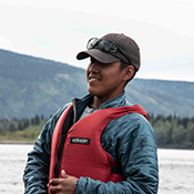 A young person wearing a life jacket and baseball cap sits in a canoe with mountains in the background. 