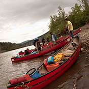 A group of paddlers dock and unload three red canoes full of supplies along the shore of a river.