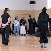A student participant carries two 45‑lb emergency assistance bags during a police training exercise in a gymnasium, while others observe.