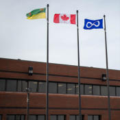 Three flags—Saskatchewan, Canada, and Métis—flying on poles in front of a brick building under an overcast sky.
