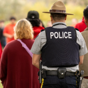 Police officer in uniform with a vest labeled POLICE stands among a group of people outdoors in red shirts.