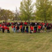 A large group of people gathered outdoors in a grassy area, forming a wide circle around a central space. Many participants are wearing red shirts or clothing, and some are dressed in patterned skirts.