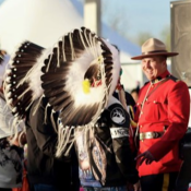An RCMP officer in a red serge uniform stands smiling near three people wearing traditional headdresses.