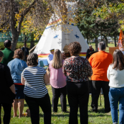 Police officer in uniform with a vest labeled POLICE stands among a group of people outdoors in front of a tipi.