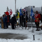 An RCMP officer in a red serge uniform salutes a number of flags in front of a snowy outdoor memorial. 