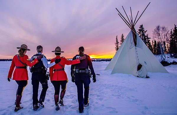 Four RCMP members walk across snow towards a tipi as the sunset
