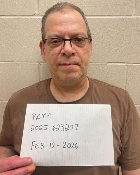 A man wearing glasses appears holding a page with identification and a file number for a police mug shot.