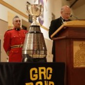Two officers in red uniforms stand behind a silver trophy while a man in a dark suit speaks at a podium. An officer in a red blazer watches from the right side of the scene.