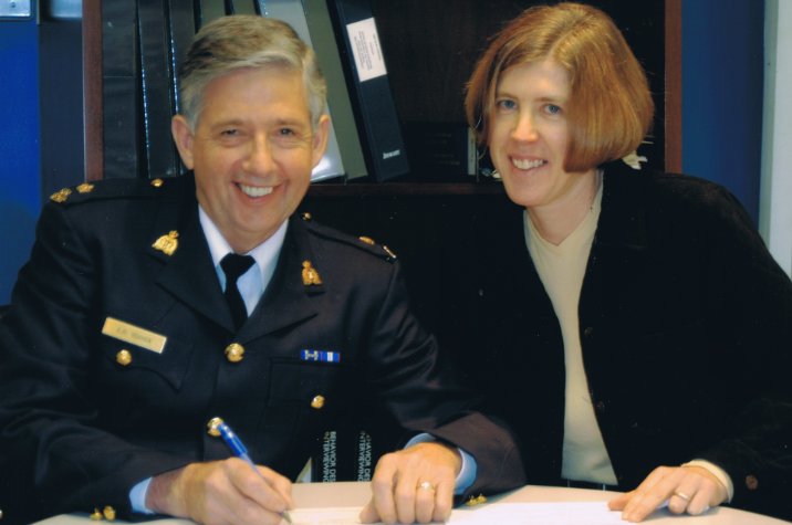 A civilian employee signs documents alongside an RCMP officer in an office setting.