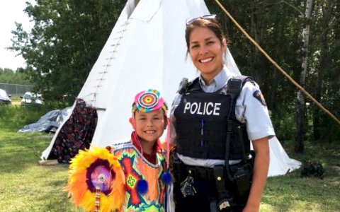 Indigenous woman police officer stands next to young boy in traditional Indigenous garb. 