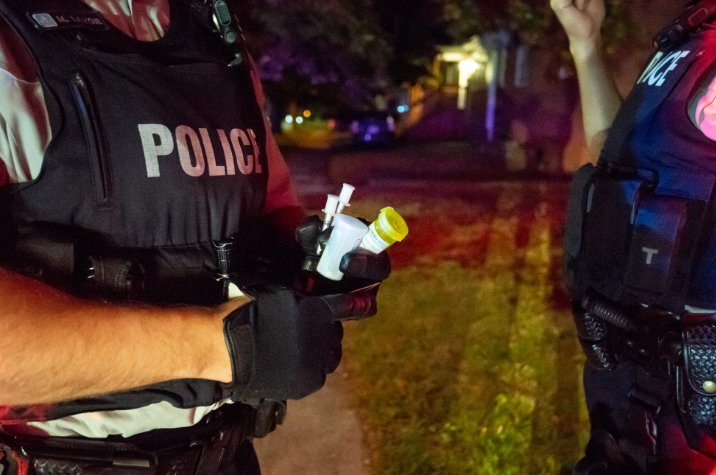 Close up of RCMP officers during a nighttime roadside interaction, with one officer holding a prescription pill bottle. Emergency lights cast red and blue reflections in the background.