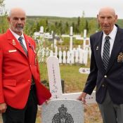 Two elderly men in blazers flank an RCMP regimental headstone amidst a field of white crosses in a cemetery.