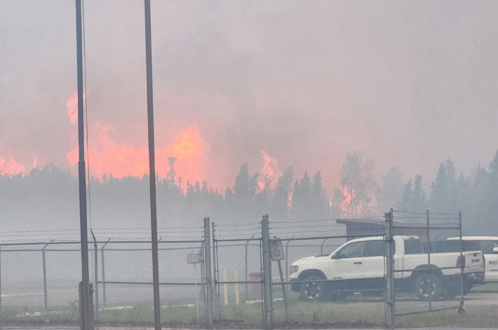 In the distance, dark orange flames burn in a wooded area, giving off dark grey smoke. In the middle ground, a gated enclosure contains two white vehicles.