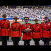 Five officers in red serge pose on a football field with a full stadium in the background. A man in the centre holds a football.