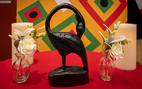 A Sankofa bird, flanked by white roses and candles, sits on a table covered with a red table cloth.