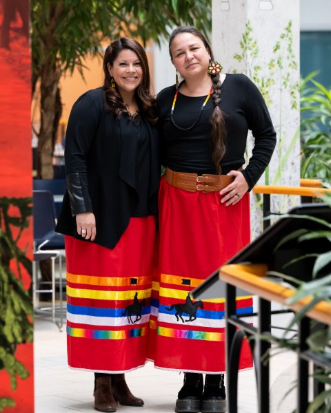 Two women dressed in black shirts and red, ankle-length skirts with the RCMP horse and rider emblem and multiple-coloured ribbons.