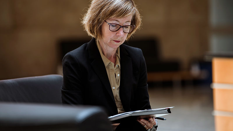 A woman in a black suit sits holding a notepad and pen