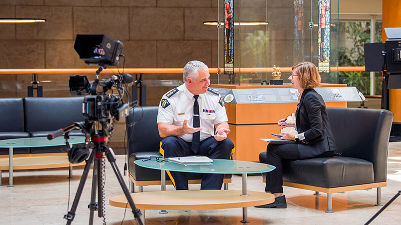 A woman and man, dressed in an RCMP uniform, sit opposite each other surrounded by cameras