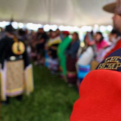The shoulder flash of an <abbr>RCMP</abbr> member in red serge is shown in front of a blurred background of a dancer at the Abegweit First Nations Pow Wow.