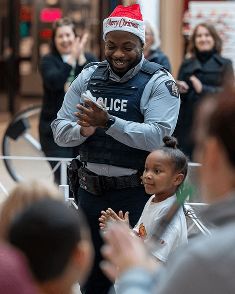 An RCMP officer in a holiday hat standing beside a child and clapping.