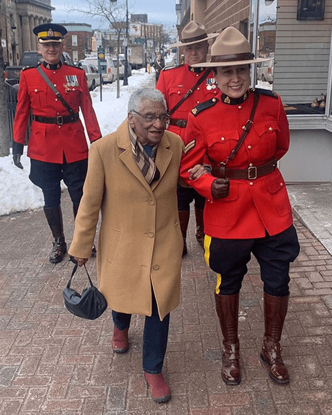 A woman walking down the street holding onto the arm of an RCMP officer. Two other RCMP officers are following behind. All officers are wearing red serge.