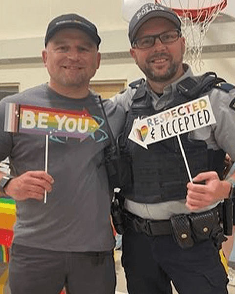 Two police officers, one in plain clothes and one in uniform, holding up signs that say, “Be you” and “Respected and accepted.”