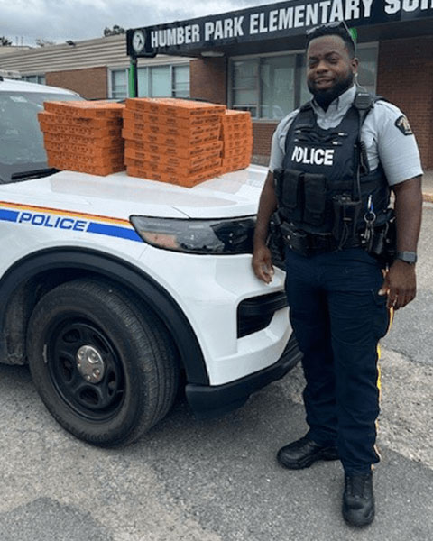 An RCMP officer standing in front of his police vehicle, which has several pizzas sitting on the hood.
