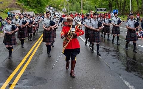 The RCMP Pipes, Drums and Dancers band walks on a wet roadway toward the camera.