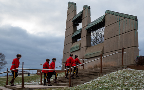 Five RCMP officers in red serge walk up stairs toward a large monument.