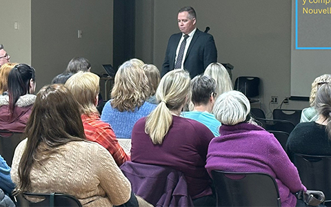 An RCMP officer in a suit stands in front of a crowded room.