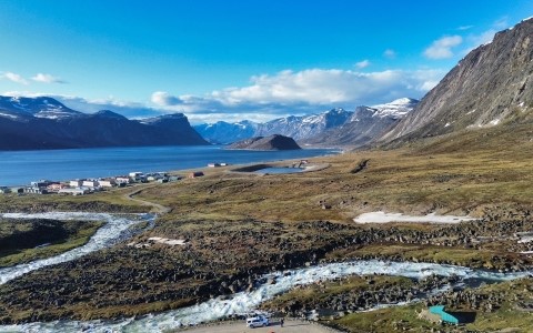 Aeriel shot of a rocky grass terrain next to a glacial lake with snow-capped mountains in the distance.