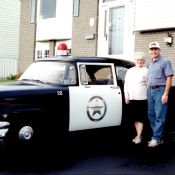 A man and an elderly woman pose outside next to a vintage police patrol car.