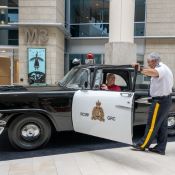An elderly man in a red shirt sits inside a vintage RCMP patrol vehicle, while a uniformed RCMP officer holds open the door. Image by Martine Chenier, RCMP