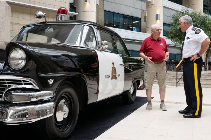 A uniformed RCMP officer and an elderly man in a red shirt stand next to a vintage vehicle inside the lobby of a building.
