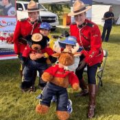 2 members in Red Serge are seen smiling with 2 children and 2 RCMP plush toys.