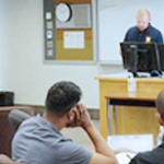 A male RCMP officer speaks to a room of new cadets in civilian clothing.