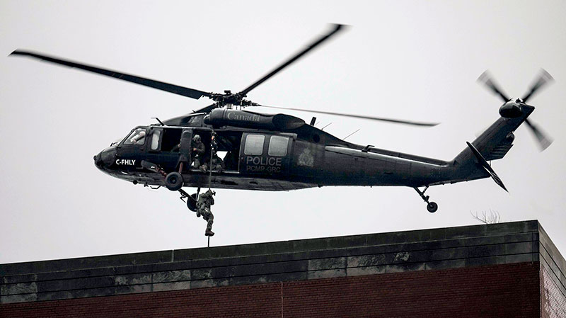 An RCMP UH-60 Black Hawk helicopter hovers above the roof of a building with a person exiting the helicopter by rope onto building’s roof.