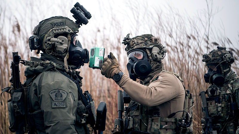 Three people in tactical gear using a using chemical, biological, radiological, and nuclear tool on an RCMP officer.