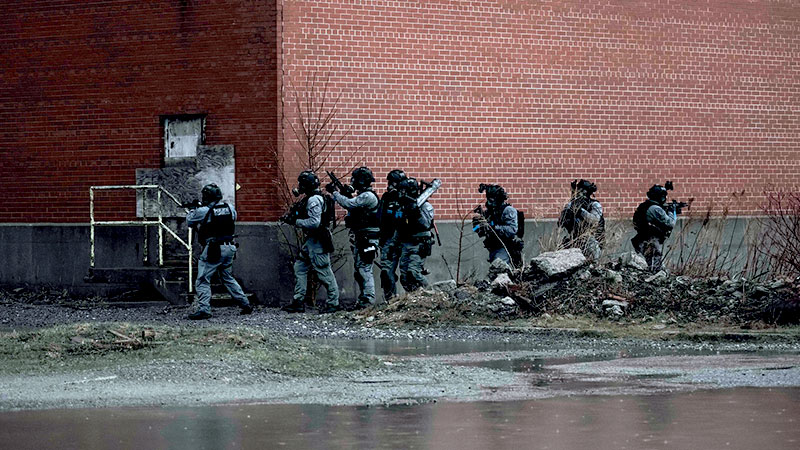 Seven people in tactical gear surround abandoned brick building.