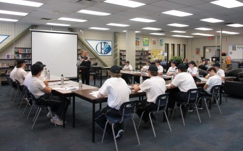 A group of teens sit at desks while a woman RCMP officer stands talking in front of them.