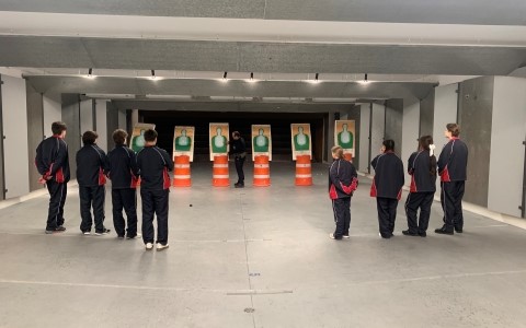 A group of teens in black uniforms stand in a row inside a firearms range.
