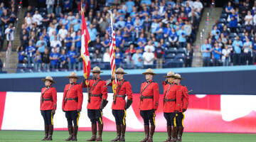 7 RCMP officers in Red Serge stand on a baseball field on a backdrop of fans in the stands.