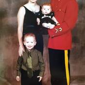 Photo of Cpl. Scotty Schumann, wearing his RCMP red serge uniform at his graduation from RCMP Training Academy. The photograph includes his wife standing beside him wearing a black dress along with his son, age 4 wearing a green shirt with a tie and black pants. Cpl. Schumann is holding his 4-month-old daughter in his arms.