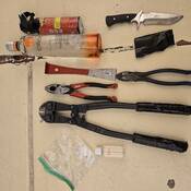 Various tools, including pliers, a bolt cutter, a snipping tool, a small pry bar, a black and silver hunting knife, a red canister of bear spray and a bottle of liquor on a yellow tile floor.